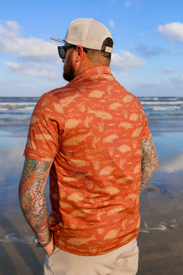 Back angled view of man wearing orange Texas Slam performance polo on the beach.