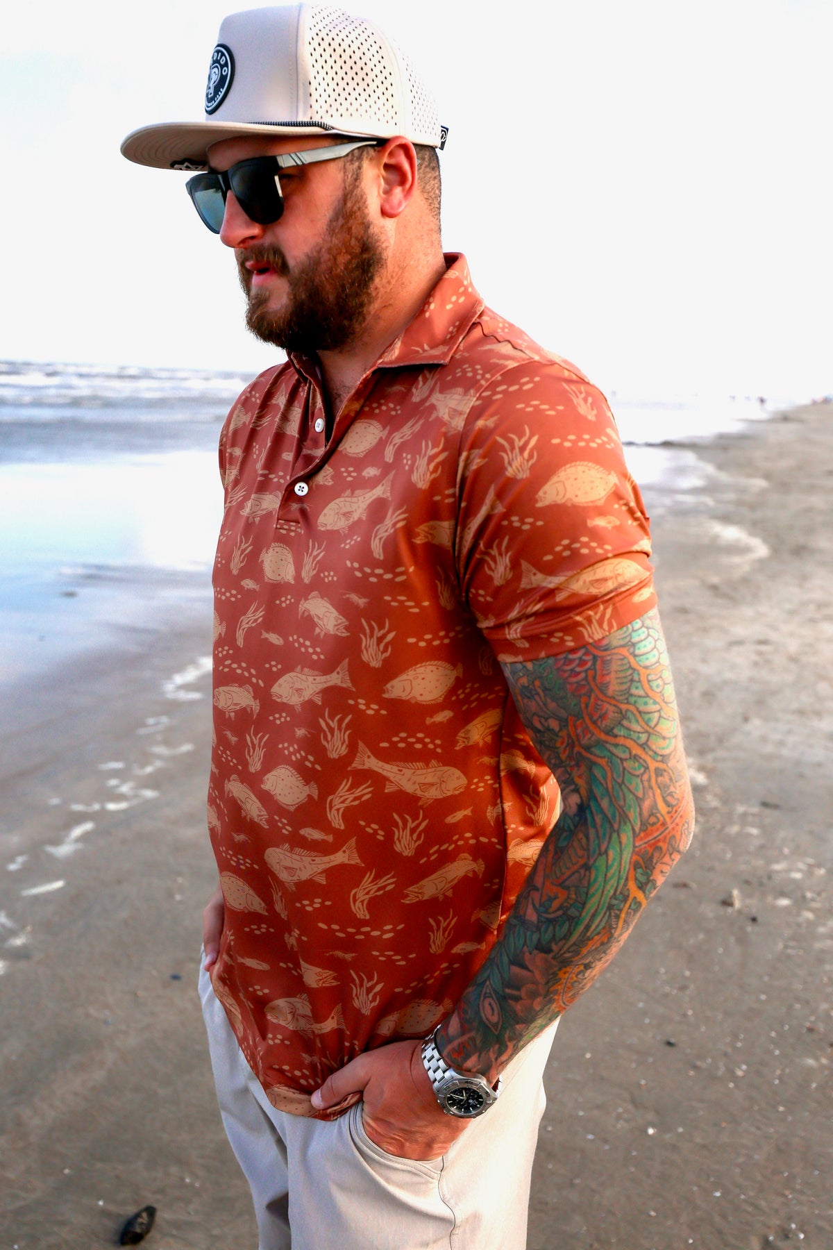 Side angled shot of man on beach wearing the patterned orange Texas Slam polo.