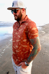 Side angled shot of man on beach wearing the patterned orange Texas Slam polo.