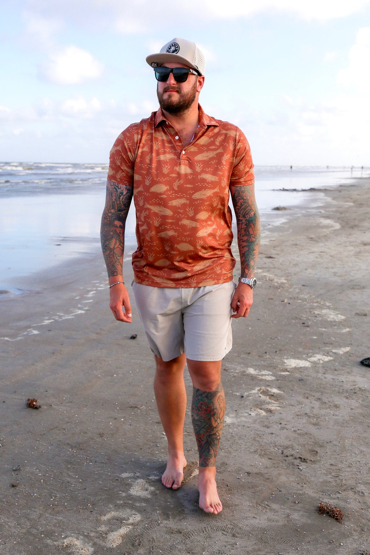 Full-body front view of man walking on beach in orange Texas Slam polo and shorts.